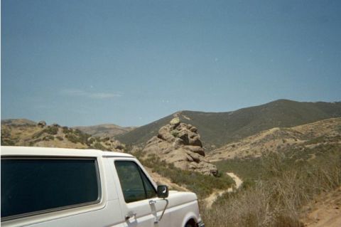 1990 Ford Bronco - front corner of a white SUV at the left edge of the frame, with a prominent rock outcrop and a winding mountain trail beyond.