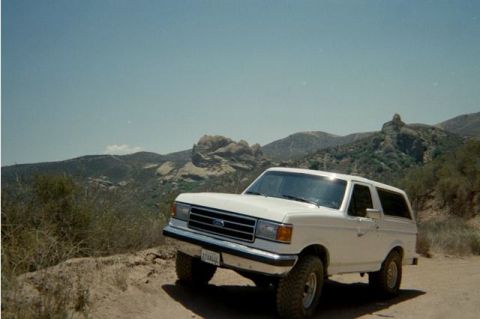 1990 Ford Bronco - white Ford Bronco parked on a sandy trail with dramatic rocky peaks rising in the background under a clear sky.