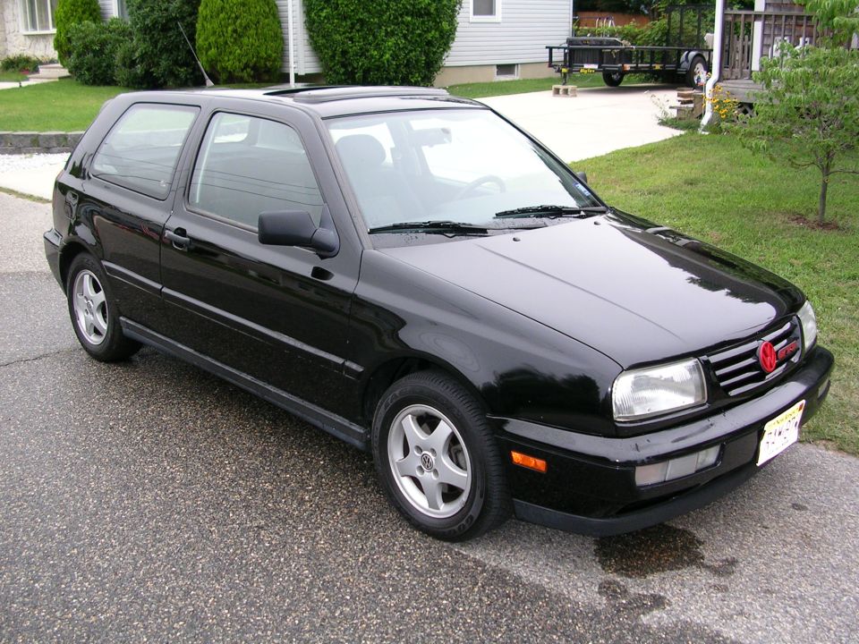 Black VW Golf 3-door hatchback, front-right 3/4 view, parked on a street near houses.