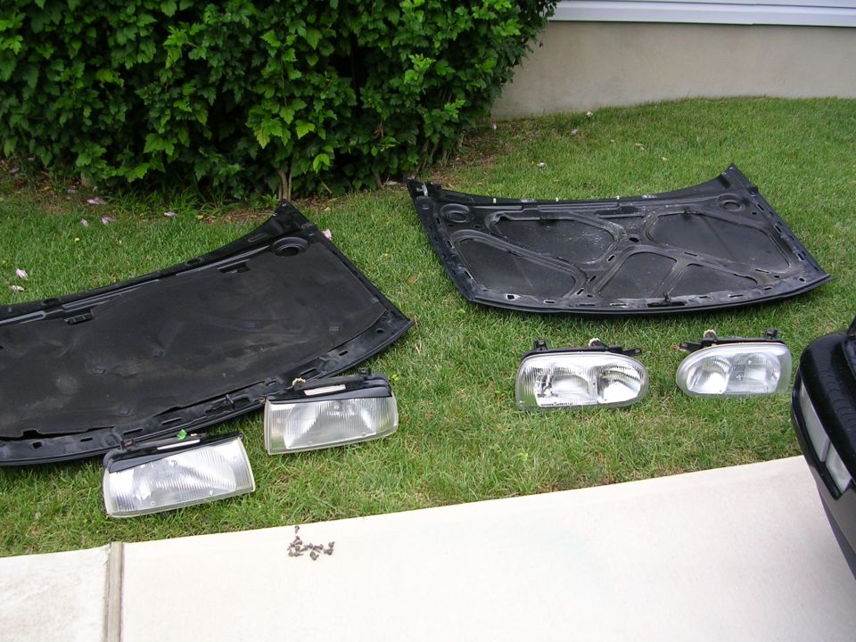 Two car hoods and a pair of headlight assemblies laid out on a grassy lawn