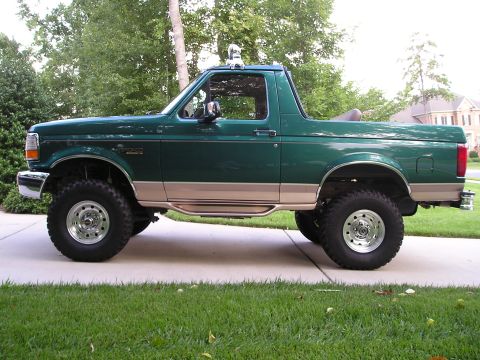 1996 Ford Bronco - This lifted green SUV is parked on a concrete driveway with grass in the foreground and trees in the background.