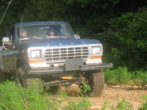 1979 Ford F-150 - A blue pickup truck drives on a dirt road surrounded by greenery, with two occupants visible.