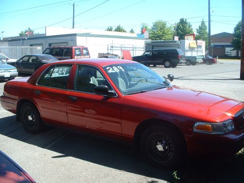 1999 Ford Crown Victoria - A red sedan is parked in a lot with other vehicles and buildings in the background.