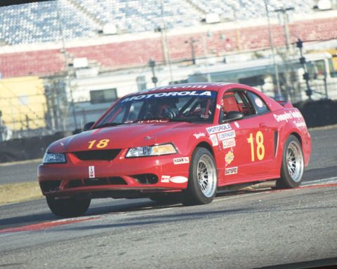 1995 Ford Mustang - A red race car with sponsor decals drives on a track, with grandstands in the background.