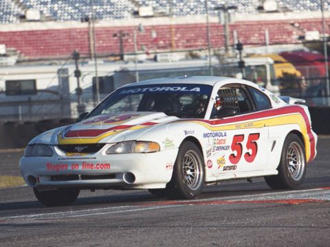 1995 Ford Mustang - A race car with sponsor decals drives on a track with grandstands in the background.