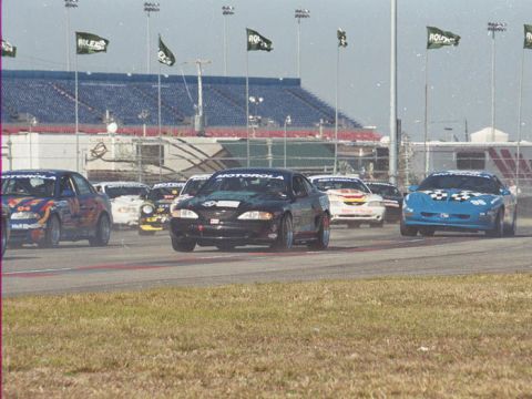 1995 Ford Mustang - Multiple race cars, including a black 1995 Ford Mustang, compete on a track with grandstands in the background.