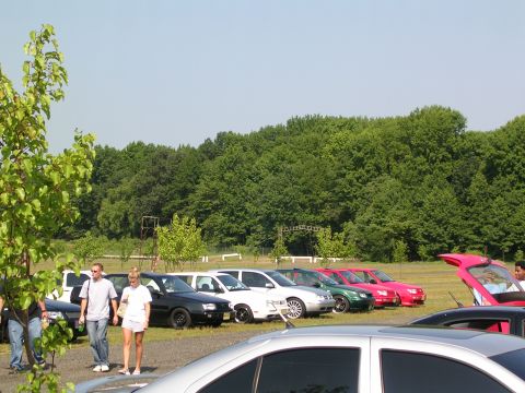 1990 Jeep YJ - A variety of cars are parked on a grassy field under a clear blue sky, with a dense line of trees forming the backdrop.