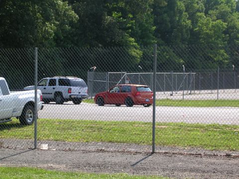 1990 Jeep YJ - An orange hatchback is parked behind a chain-link fence, with other vehicles visible in the background.