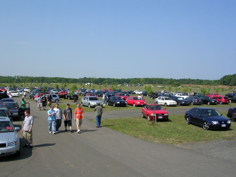 Outdoor car show with many cars parked on grass and asphalt under a clear blue sky