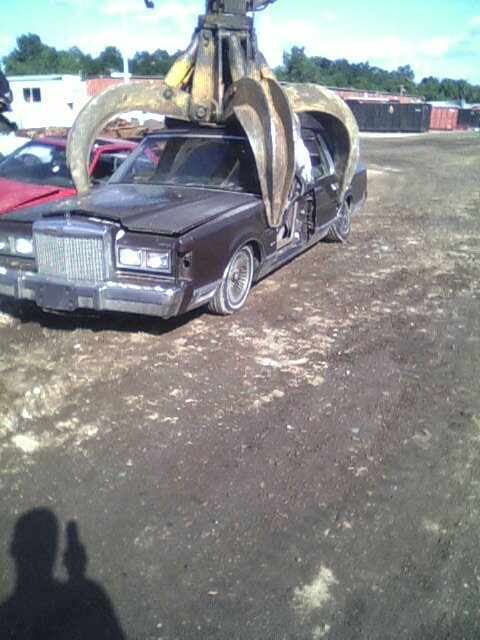 1986 Lincoln Town Car - A 1986 Lincoln Town Car is lifted by a claw crane at a junkyard, with another vehicle visible in the background.