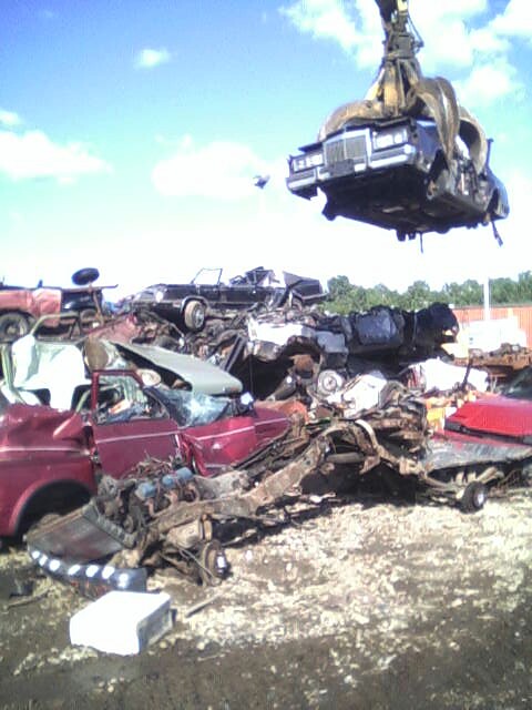 1986 Lincoln Town Car - Crane lifts a 1986 Lincoln Town Car over a junkyard filled with wrecked vehicles.