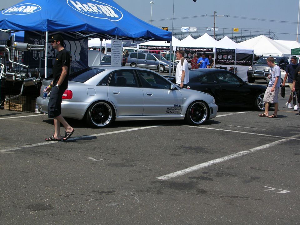 Silver sedan with black aftermarket wheels parked at an outdoor event, another black car behind it