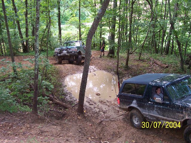 1995 Ford Bronco and a green off-road truck on a muddy path in the woods