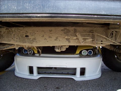 1989 Ford Bronco - The front of a yellow car with custom headlights is tucked under a larger vehicle's chassis.