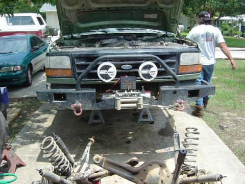 1993 Ford Bronco - A man works on a green Ford F-Series truck that is raised on jack stands with suspension components laid out.
