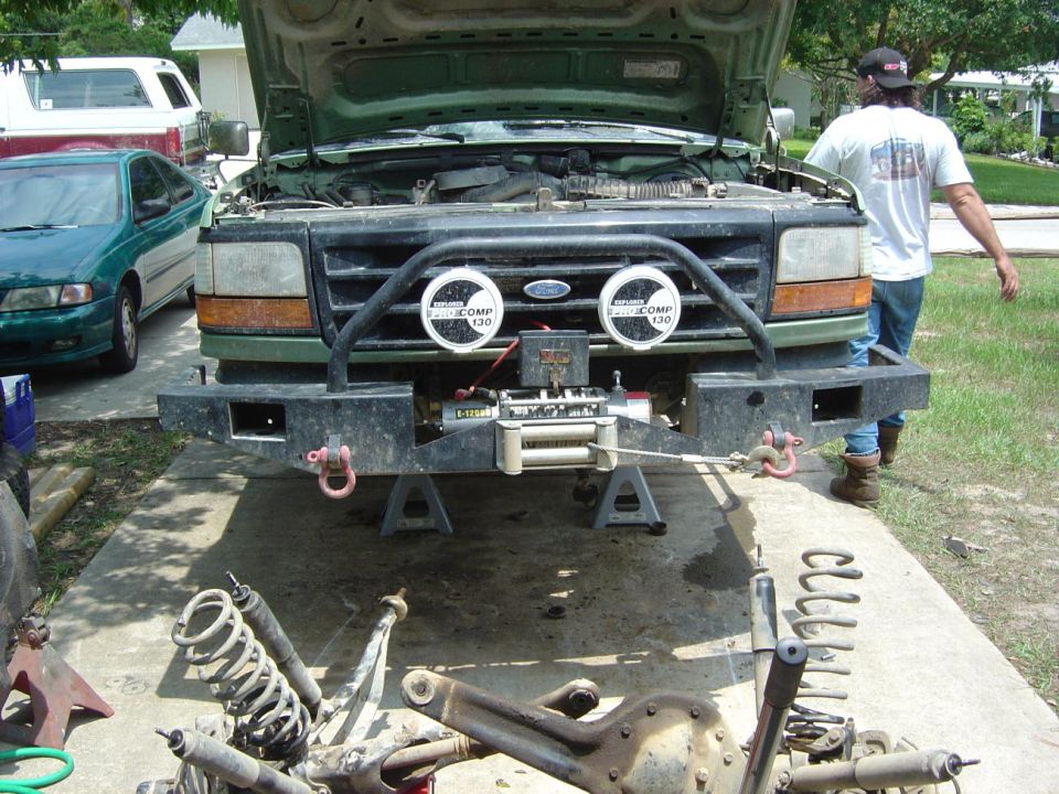 Green Ford F-Series truck with hood up, winch on bumper, and suspension parts on ground. Another car and van in background