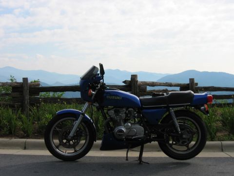 1985 Ford LTD Crown Victoria - A blue Suzuki motorcycle is parked by a wooden fence with mountains in the distance.