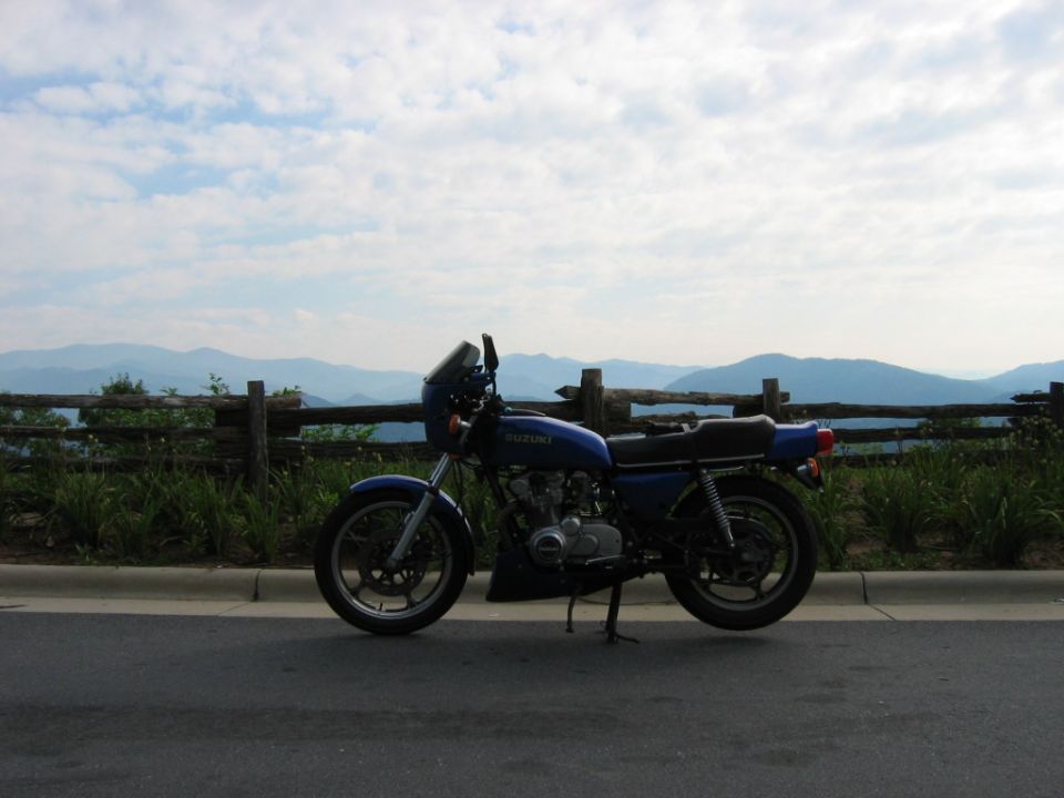 Blue Suzuki motorcycle parked on the side of a road with a wooden fence and mountains in the background
