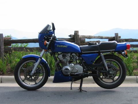 1985 Ford LTD Crown Victoria - A blue Suzuki motorcycle is parked on the side of a road with a wooden fence and mountains visible behind it.