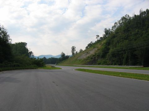 1985 Ford LTD Crown Victoria - A two-lane road winds through a scenic landscape with a steep, tree-covered hill on the right.