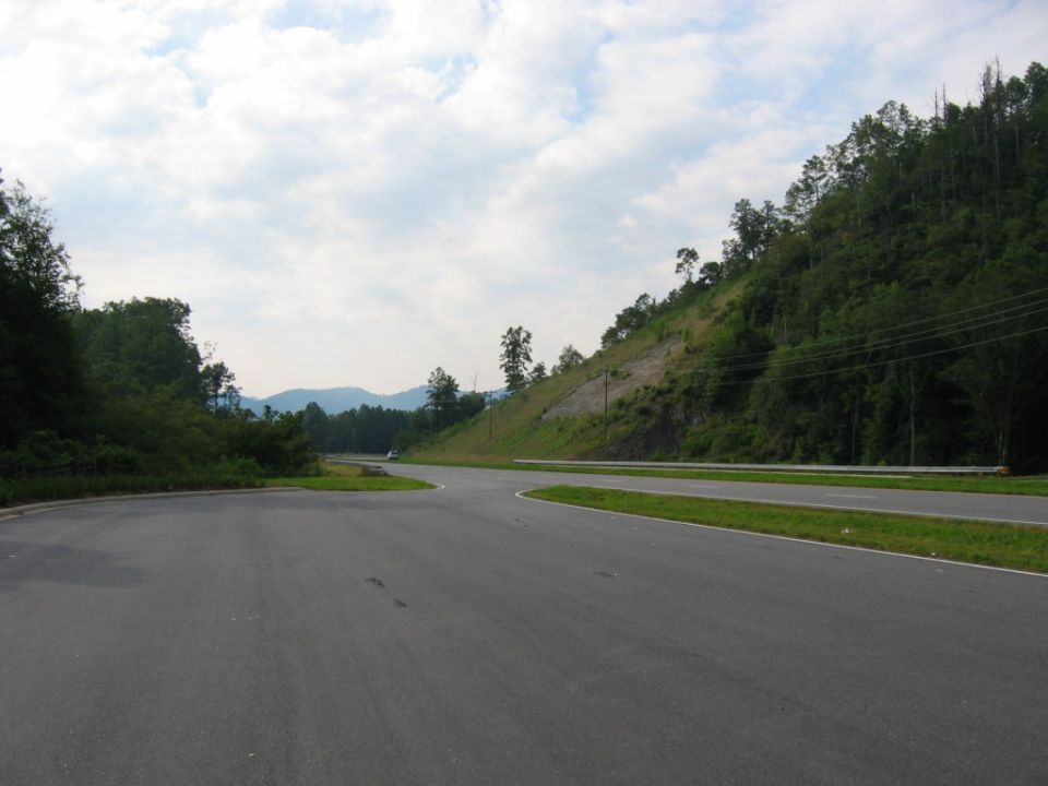 Roadway curves through a hilly, wooded area under a cloudy sky. A distant car is visible
