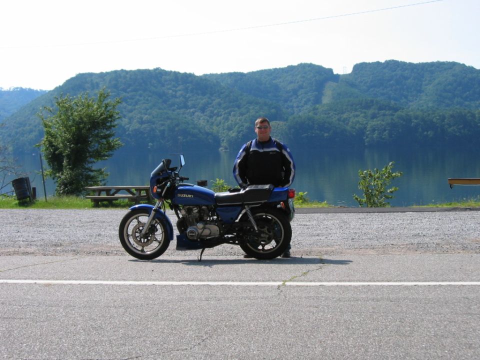 Blue Suzuki motorcycle parked on asphalt with a rider standing next to it; lake and mountains in background