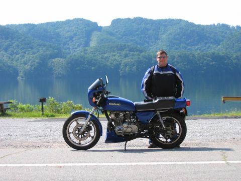 1985 Ford LTD Crown Victoria - Man and his blue Suzuki motorcycle parked by a lake with mountains in the distance.