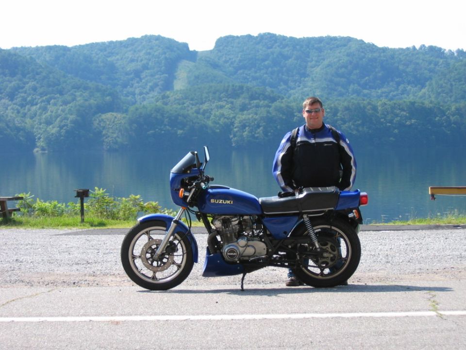 Man in motorcycle gear stands next to a blue Suzuki motorcycle. Lake and mountains in background