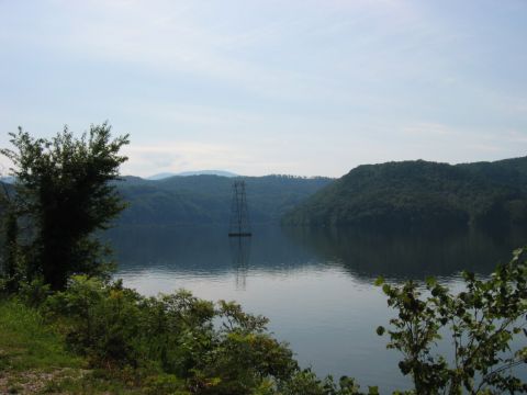 1985 Ford LTD Crown Victoria - A solitary power line tower stands in the middle of a tranquil lake, bordered by lush green hills.