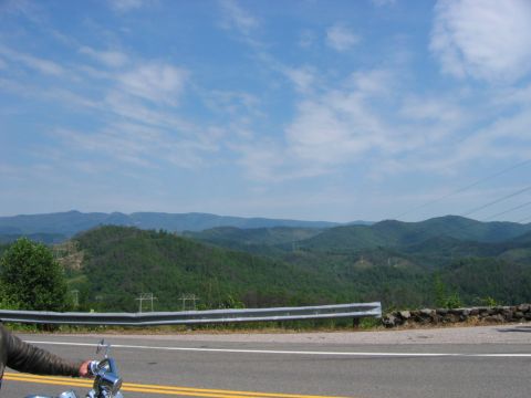 1985 Ford LTD Crown Victoria - A motorcycle rider's perspective on a scenic mountain road, with lush green hills stretching to the horizon under a partly cloudy sky.