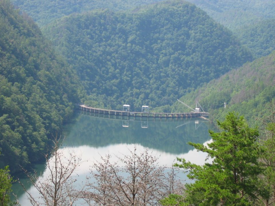 Dam structure across a reservoir with reflections, surrounded by forested mountains