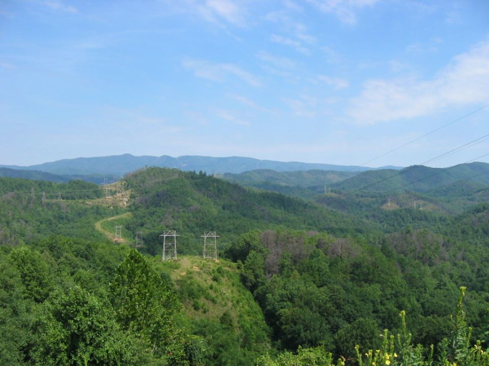 Mountainous terrain with green trees and power line towers under a blue sky