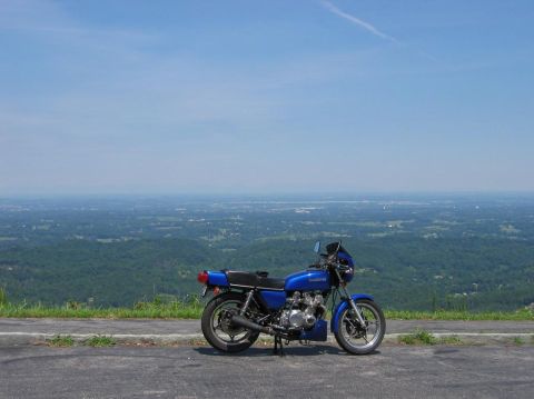 1985 Ford LTD Crown Victoria - A blue motorcycle is parked on the side of a road overlooking a vast, green valley under a clear blue sky.