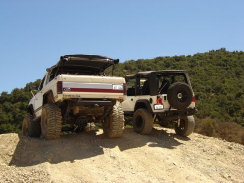 1991 Ford Bronco - Two off-road vehicles, a Ford Bronco and a Jeep Wrangler, are positioned on a dusty incline under a clear blue sky.