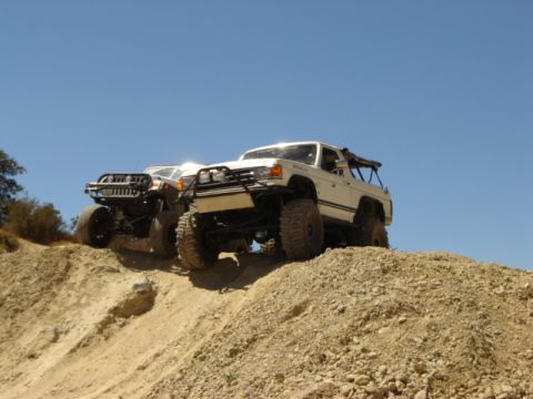 1991 Ford Bronco - Two modified SUVs ascend a steep dirt incline under a clear blue sky.