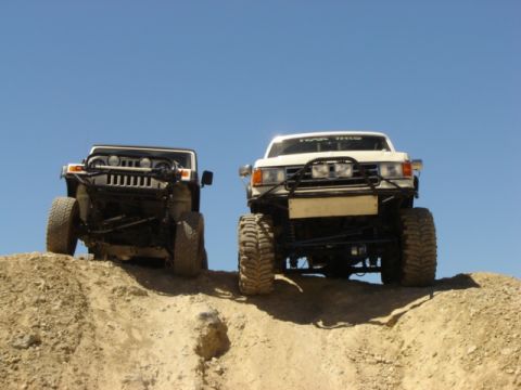 1991 Ford Bronco - Two modified trucks ascend a dirt incline, their off-road capabilities under a bright sky.