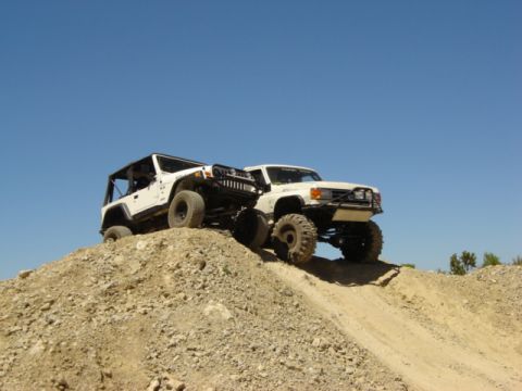 1991 Ford Bronco - Two modified SUVs navigate a steep dirt incline under a clear blue sky.