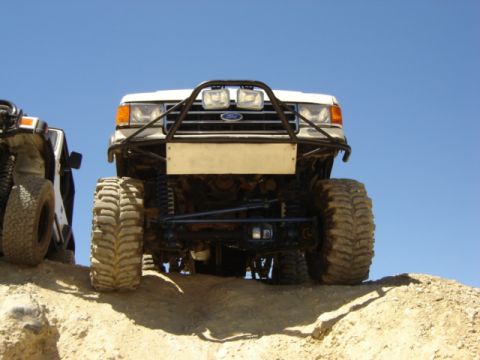 1991 Ford Bronco - Two off-road vehicles are positioned on a dirt incline against a clear blue sky.