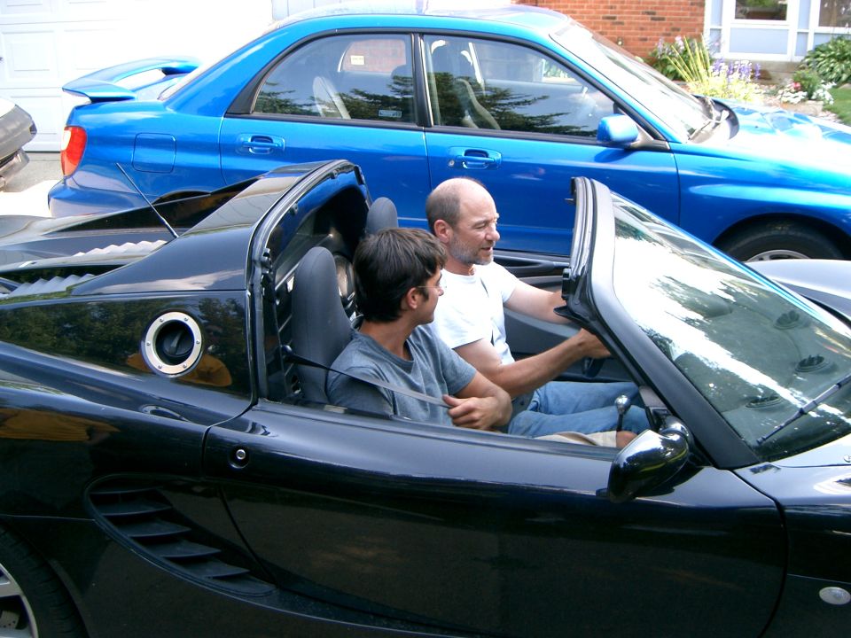 Two men seated in a black convertible roadster with a blue Subaru WRX sedan parked behind in a driveway