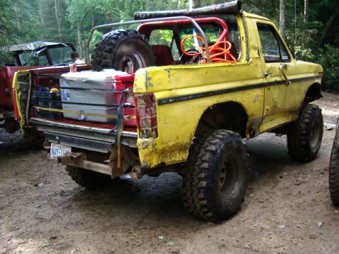 1985 Ford Bronco - Mud-splattered yellow full-size Ford Bronco with the hardtop removed, a red roll cage, exhaust stack and a rear-mounted spare tire, seen fr…