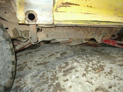 1985 Ford Bronco - Low-angle underbody shot along the rocker of a yellow vehicle in a garage, showing a rusty frame rail, body mounts and a black-capped hole…