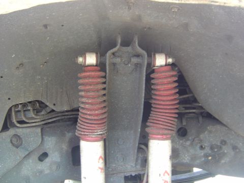 1993 Ford Bronco - Looking up at the front underside of a truck showing a pair of white-bodied shocks with red coil-over-style dust boots mounted to the cross…