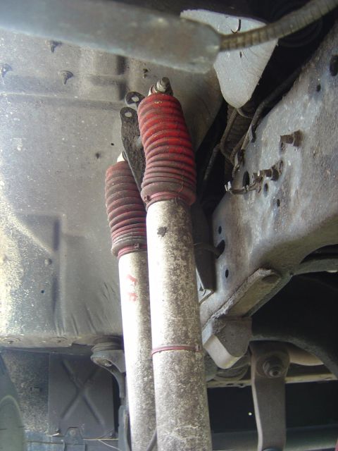 1993 Ford Bronco - Underbody view of a pair of dual shock absorbers with white bodies and red accordion boots mounted side by side to a truck frame crossmembe…