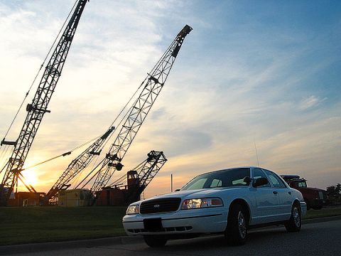 2000 Ford Crown Victoria - A white 2000 Ford Crown Victoria is parked near a construction site with large cranes silhouetted against the sunset sky.