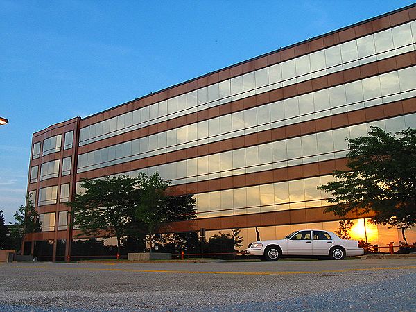 White sedan parked in front of a large office building with reflective windows. Sunset visible in the reflection