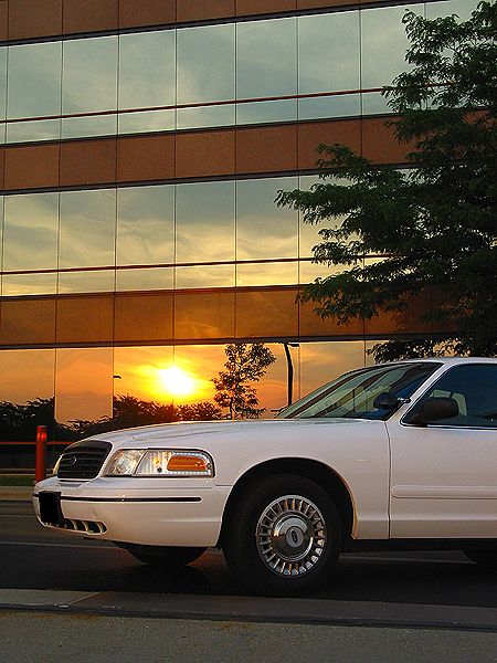 2000 Ford Crown Victoria - A white 2000 Ford Crown Victoria is parked in front of a building whose windows reflect a vibrant sunset.