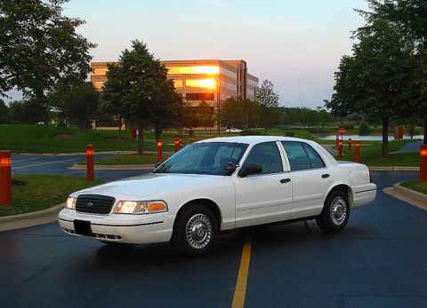 2000 Ford Crown Victoria - A white 2000 Ford Crown Victoria is parked in a lot with a modern building in the background during sunset.