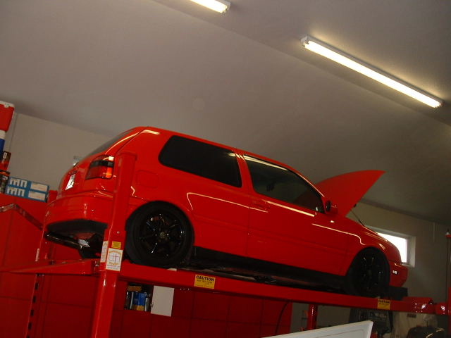 Red two-door hatchback on a shop lift with hood open and black wheels in a garage