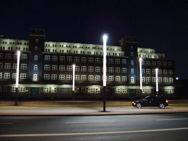 Dark car parked at night in front of a large lit brick building behind bright vertical light poles