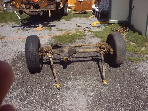 1988 Ford Bronco - A detached front axle assembly with tires sits on a gravel surface, with the rear of a vehicle in the background.